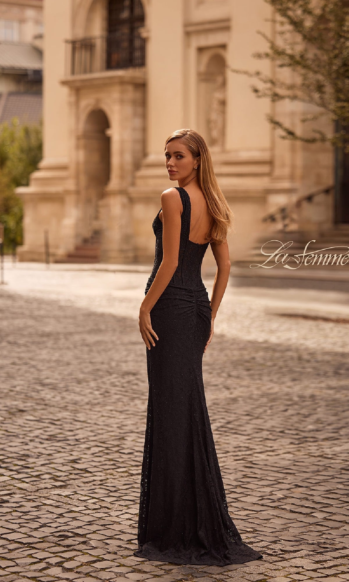 A woman in a Beaded Long Lace Prom Dress: La Femme 32943 stands on a cobblestone street, glancing over her shoulder with an elegant building in the background.